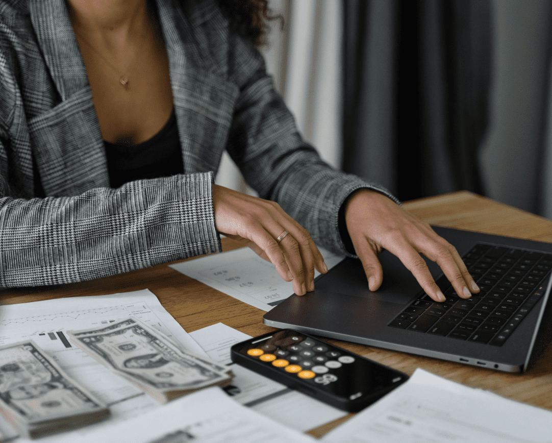 A woman uses a laptop an calculator with paperwork and piles of bills next to her on table